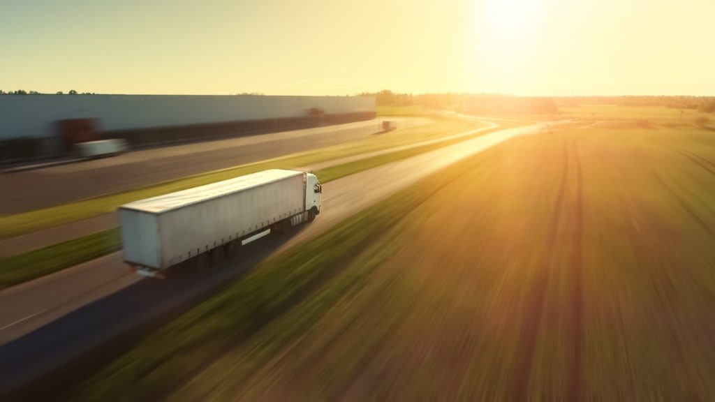 An aerial photograph of a 16-wheeler truck driving down a long stretch of road.