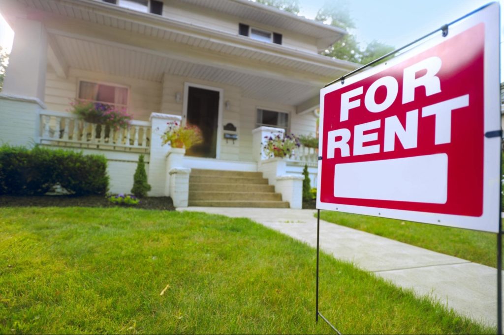 A picture of a "For Rent" sign in front of a house with a nicely manicured front lawn.