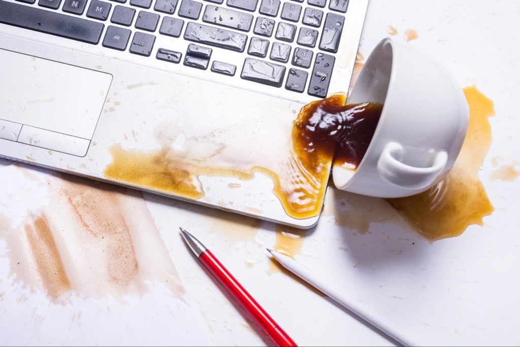 A picture of a white coffee mug tipped over with spilled coffee on the keyboard of a laptop.