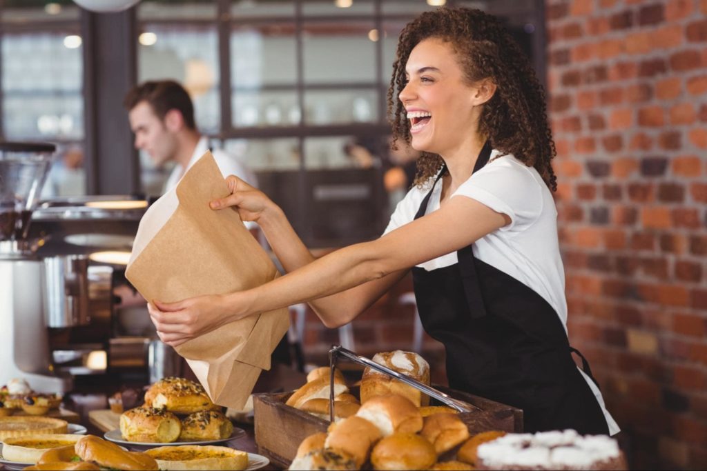A young, happy female cafe worker handing a brown paper bag filled with pastries to a customer.