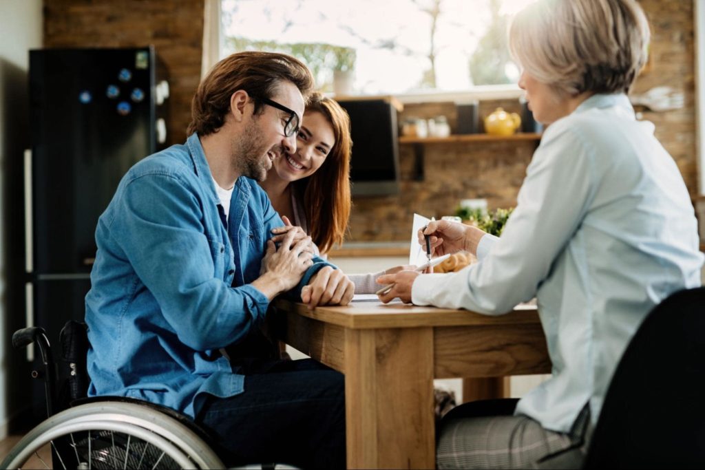 A young couple - the man is in a wheel chair and the woman is sitting next to him - talking to an insurance professional.