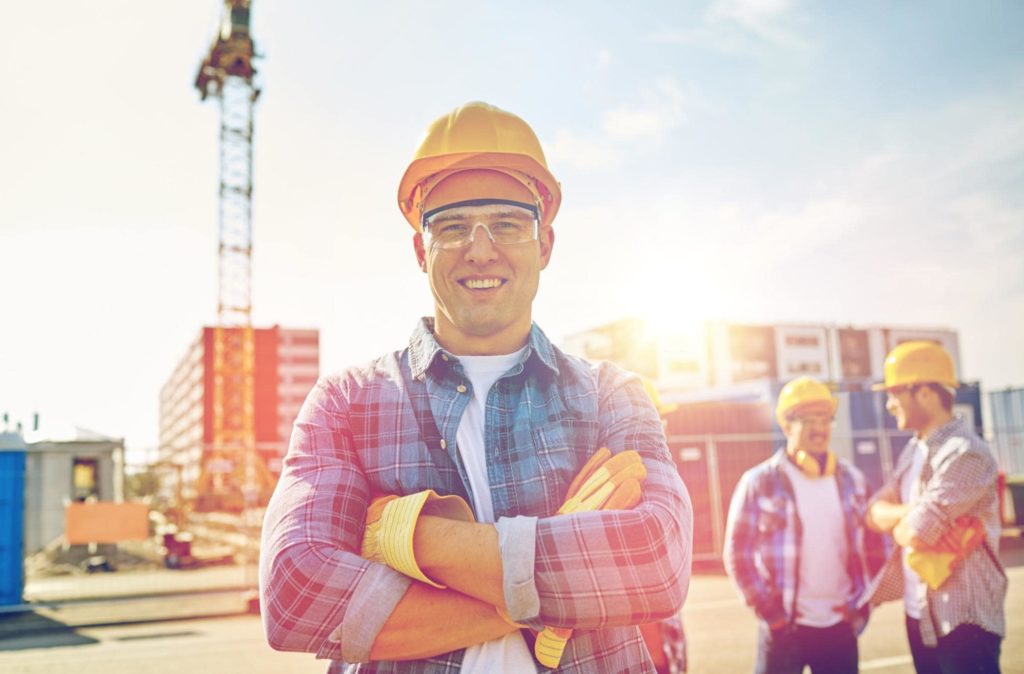 A construction worker wearing a flannel smiles with his arms crossed while two other workers talk in the background.