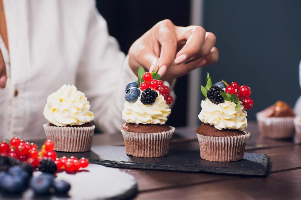 A catering employee decorating a row of three cupcakes with fruit.
