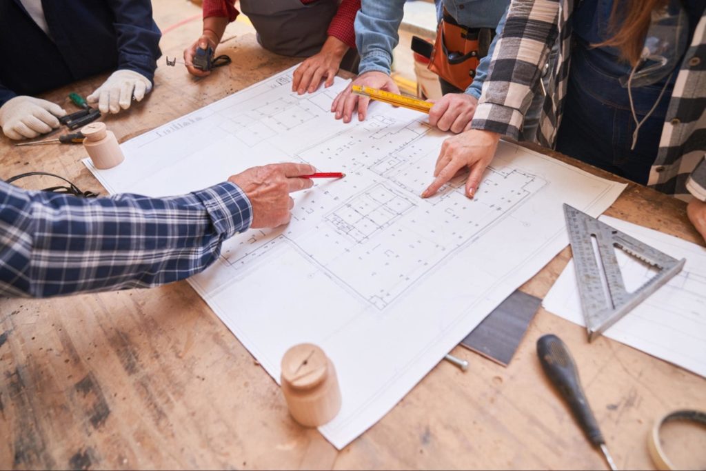 A group of construction workers around a wooden table surveying a building blueprint.